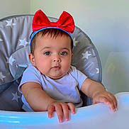 Ayzal is registered to the contest to win money with this photo: baby, high_chair, red_bow, headband, infant, child, face, hands, expression, clothing, indoor, furniture, star_pattern, person, portrait, cute, baby_girl, sitting, table, feeding_chair