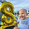 baby, smiling, balloon, number_8, golden_balloon, crown_balloon, blue_sky, outdoor, onesie, happy, child, portrait, hand, grass, cross, cemetery, reflection, car, person, celebration