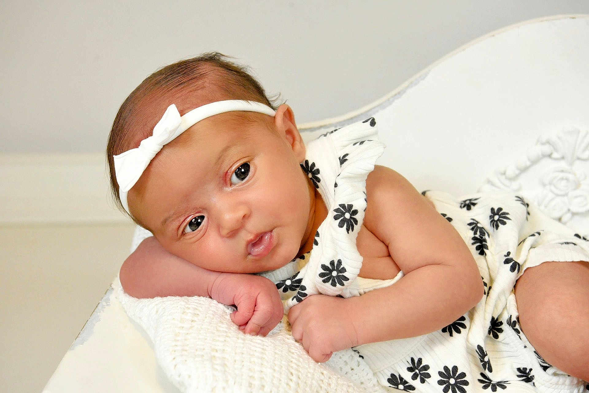 Imani joined the competition — help win amazing prizes! baby, infant, headband, white_bow, white_blanket, black_flowers, white_dress, cute, portrait, closeup, soft_texture, resting, indoors, child, newborn, skin, expression, face, hand, curious