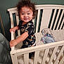 toddler, child, crib, smile, curly_hair, bedroom, blanket, furniture, indoor, person, clothing, happy, baby, standing, wood, pajamas, nursery, home, cute, infant