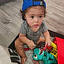 Jaxon is registered to the contest to win money with this photo: toddler, child, blue_cap, toy_phone, green_dinosaur_toy, playpen, pillow, mesh_net, curious, indoor, sitting, gray_shirt, barefoot, plastic_toys, colorful, young_child, cute, infant, playtime, home