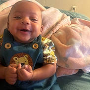Sekai is registered to the contest to win money with this photo: baby, smiling, child, overalls, lion_design, blanket, pink_blanket, blue_overalls, happy, cute, infant, portrait, indoors, person, fabric, clothing, sitting, skin, hand, face