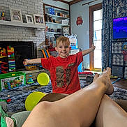 Eli joined the competition — help win amazing prizes! child, boy, red_shirt, smiling, living_room, toys, balloons, carpet, fireplace, bookshelf, curtains, window, legs, feet, person, home_interior, furniture, relaxing, play, happy