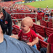 Zephaniah is registered to the contest to win money with this photo: toddler, child, adult, stadium, baseball, sports_event, crowd, seating, red_shirt, casual_clothing, outdoor, daytime, smiling, happy, people, spectators, field, fence, sports_fan, family