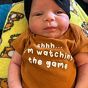 Milani is registered to the contest to win money with this photo: baby, infant, headwrap, brown_shirt, text_on_clothing, blanket, yellow_blanket, teddy_bear_pattern, hand, face, person, indoor, cute, resting, calm, clothing, newborn, portrait, child, baby_girl