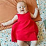 baby, infant, red_dress, smiling, lying_down, blanket, cushion, happy, child, cute, indoors, pale_skin, patterned_fabric, small_hands, small_feet, cheerful, portrait, head, person, soft_light
