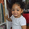child, toddler, baby, portrait, smiling, curly_hair, white_shirt, standing, indoors, bookshelf, vinyl_records, music, home, living_room, furniture, shelf, red_bag, dark_skin, face, candid