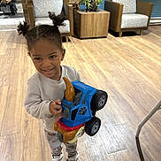 Kadrian is registered to the contest to win money with this photo: chair, child, floor, furniture, hair, happy, indoor, pants, person, pigtails, plant, playful, shoes, smile, sweatshirt, toddler, toy, truck, wooden_floor, young