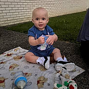 Elliott is registered to the contest to win money with this photo: baby, child, blanket, toy, bottle, outdoor, grass, brick_wall, sitting, footwear, shirt, shorts, smiling, cute, person, daylight, sidewalk, playtime, young_child, curious