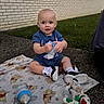 baby, child, blanket, toy, bottle, outdoor, grass, brick_wall, sitting, footwear, shirt, shorts, smiling, cute, person, daylight, sidewalk, playtime, young_child, curious