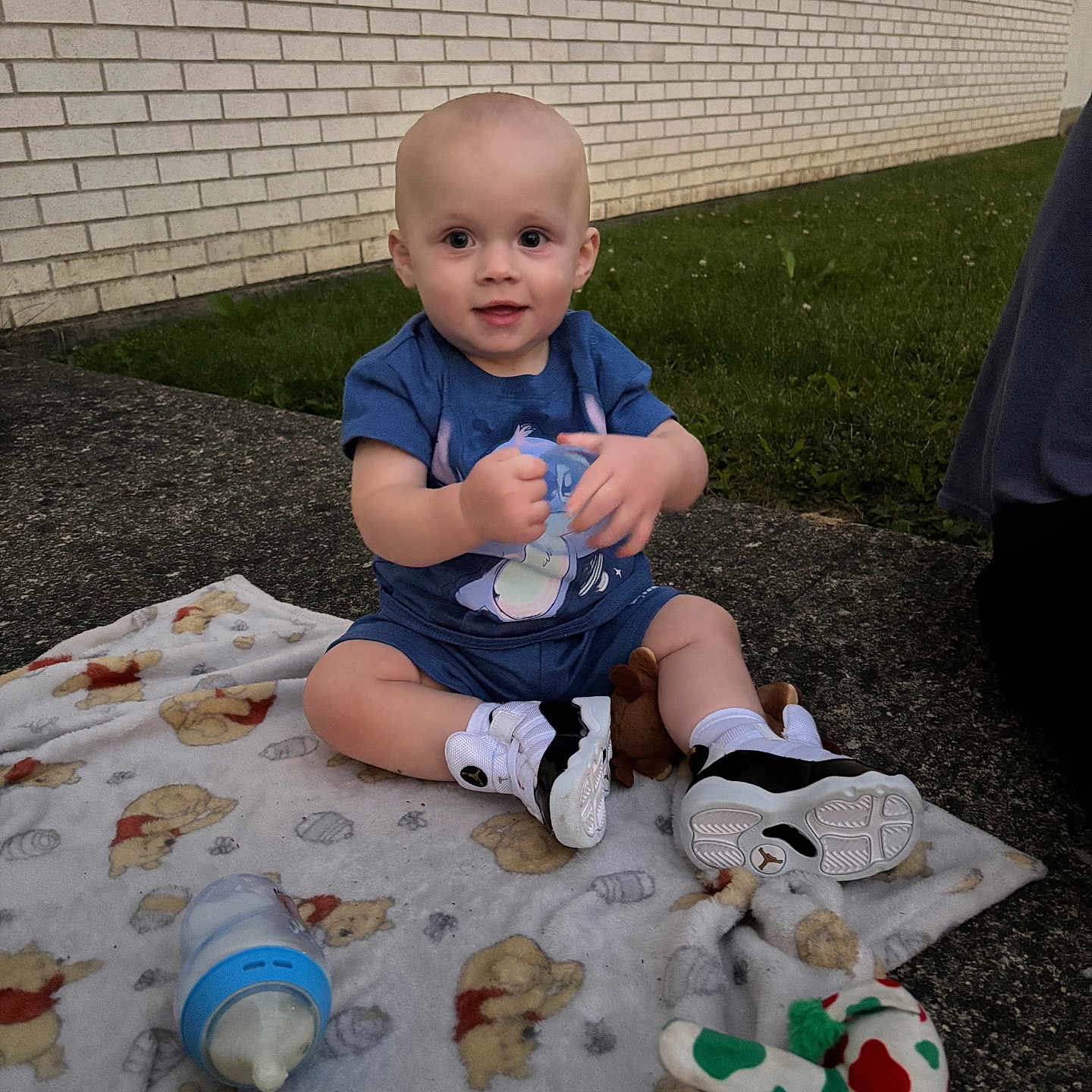 Elliott is registered to the contest to win money with this photo: baby, child, blanket, toy, bottle, outdoor, grass, brick_wall, sitting, footwear, shirt, shorts, smiling, cute, person, daylight, sidewalk, playtime, young_child, curious
