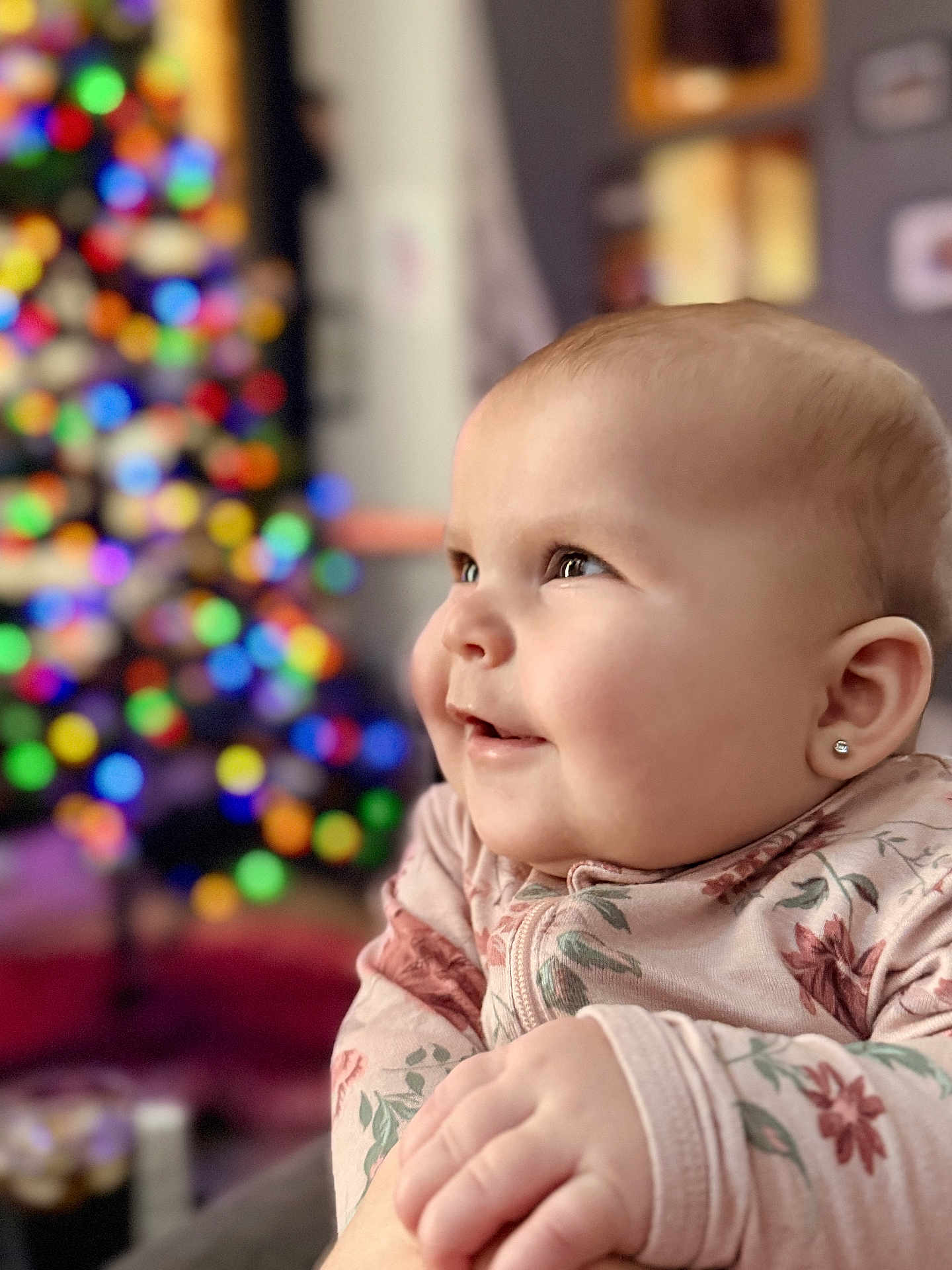 Gemma is registered to the contest to win money with this photo: baby, child, face, earring, smile, happy, indoor, portrait, christmas_lights, blurred_background, holiday, cute, soft_light, infant, clothing, long_sleeve, pink, floral_pattern, hand, closeup