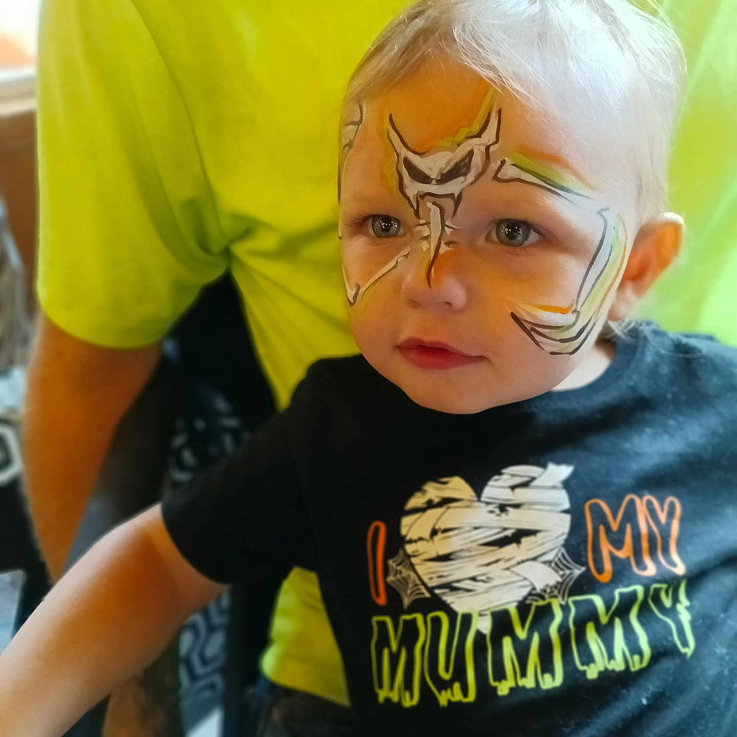 Ranger is registered to the contest to win money with this photo: black_shirt, blond_hair, blue_eyes, casual, child, closeup, costume, cute, expression, face_paint, halloween, indoor, parent, person, portrait, sitting, tattooed_arm, text_on_shirt, toddler, yellow_shirt