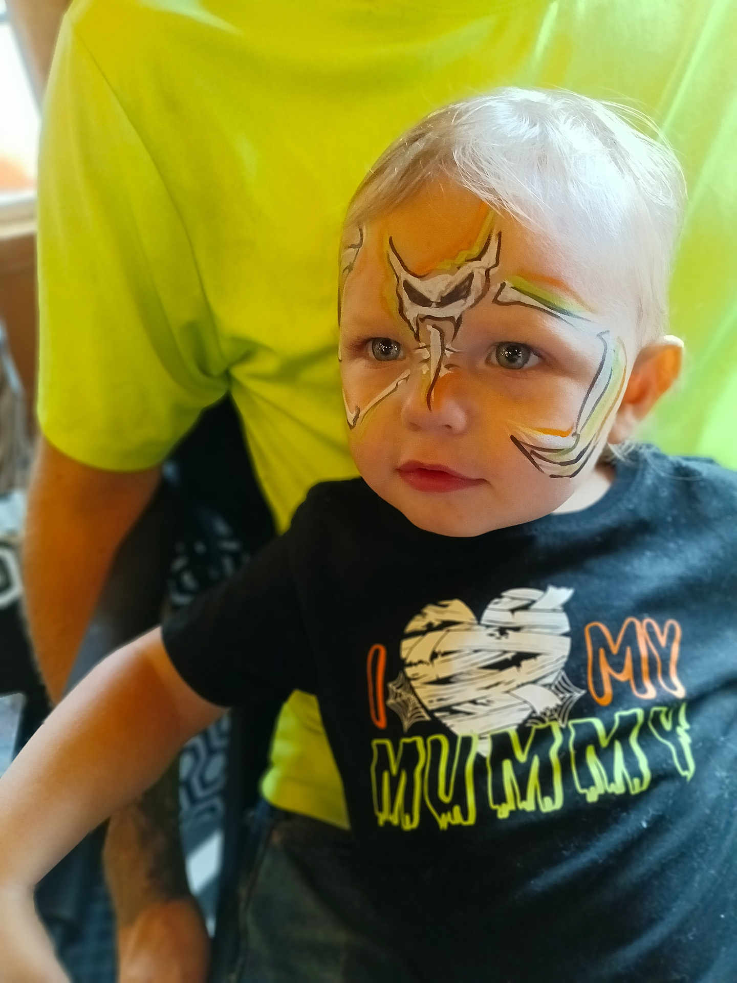 Ranger is registered to the contest to win money with this photo: toddler, child, face_paint, portrait, black_shirt, text_on_shirt, person, indoor, sitting, cute, blond_hair, blue_eyes, tattooed_arm, yellow_shirt, parent, closeup, expression, costume, halloween, casual