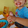 child, toddler, smiling, eating, pasta, noodles, tomato_sauce, fork, food, messy_face, blue_shirt, short_hair, table, chair, napkin, bowl, indoor, portrait, happy, playful