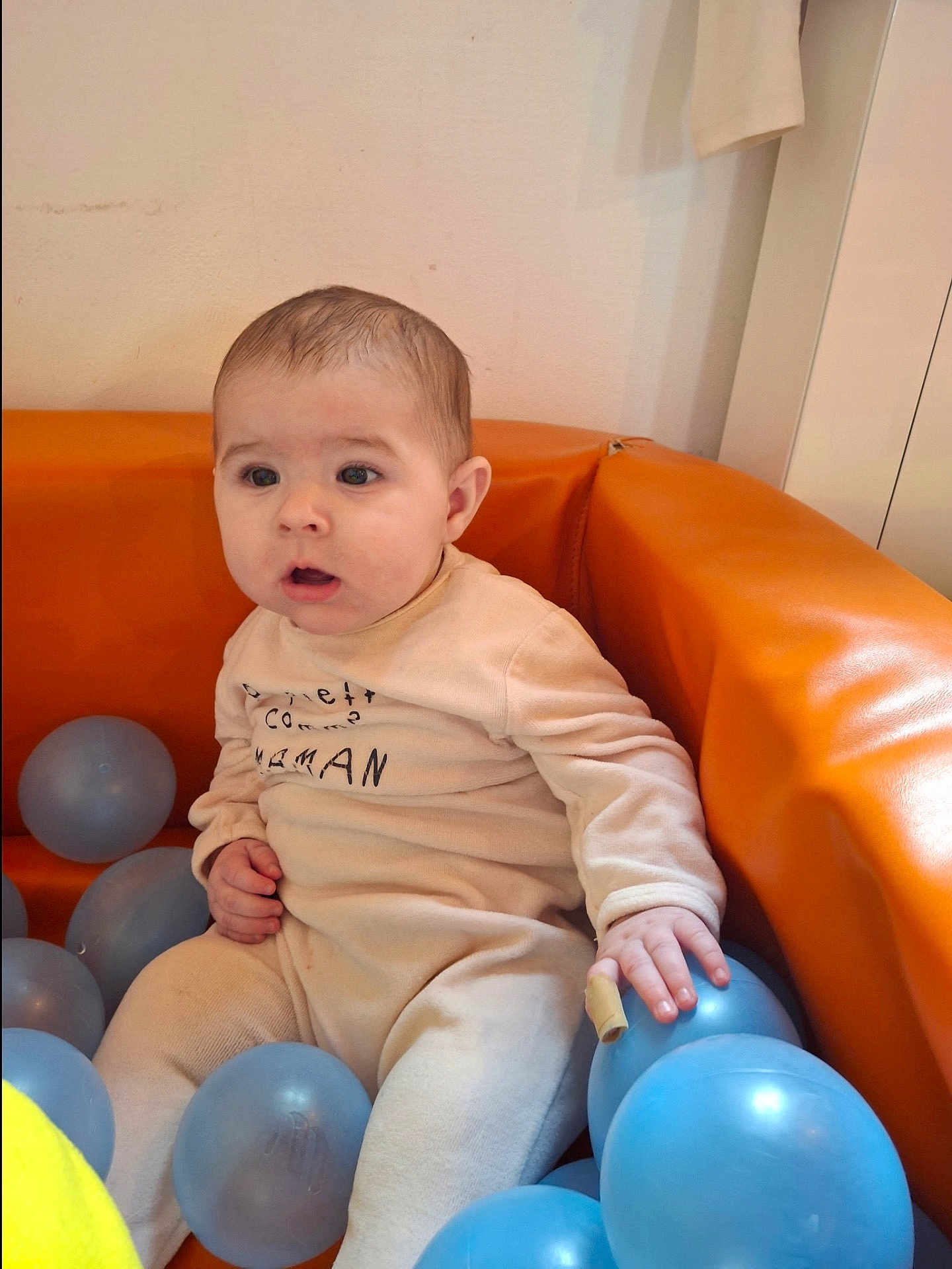 Noa a rejoint le concours — aidez-le/la à gagner de superbes lots ! baby, infant, child, sitting, orange_sofa, ball_pit, blue_ball, balls, onesie, clothing, hand, face, eyes, surprised_expression, indoor, wall, play_area, portrait, cute, soft_furniture