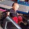 baby, bed, bedroom, black_car, blanket, carpet, child, clothing, fan, furniture, harness, home, indoor, infant, playpen, portrait, red_pants, ride_on_car, seated, toy_car