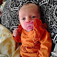 Amelia is registered to the contest to win money with this photo: baby, infant, pacifier, orange_clothing, lace, patterned_pillow, blanket, bed, hand, face, eyes, cute, lying_down, indoor, comfort, soft_fabric, newborn, child, resting, portrait