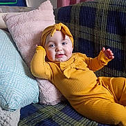 Amelia is registered to the contest to win money with this photo: baby, child, infant, smile, yellow_outfit, headband, couch, pillow, plaid, cozy, indoor, relaxing, cute, portrait, person, furniture, home, soft_texture, happy, comfort