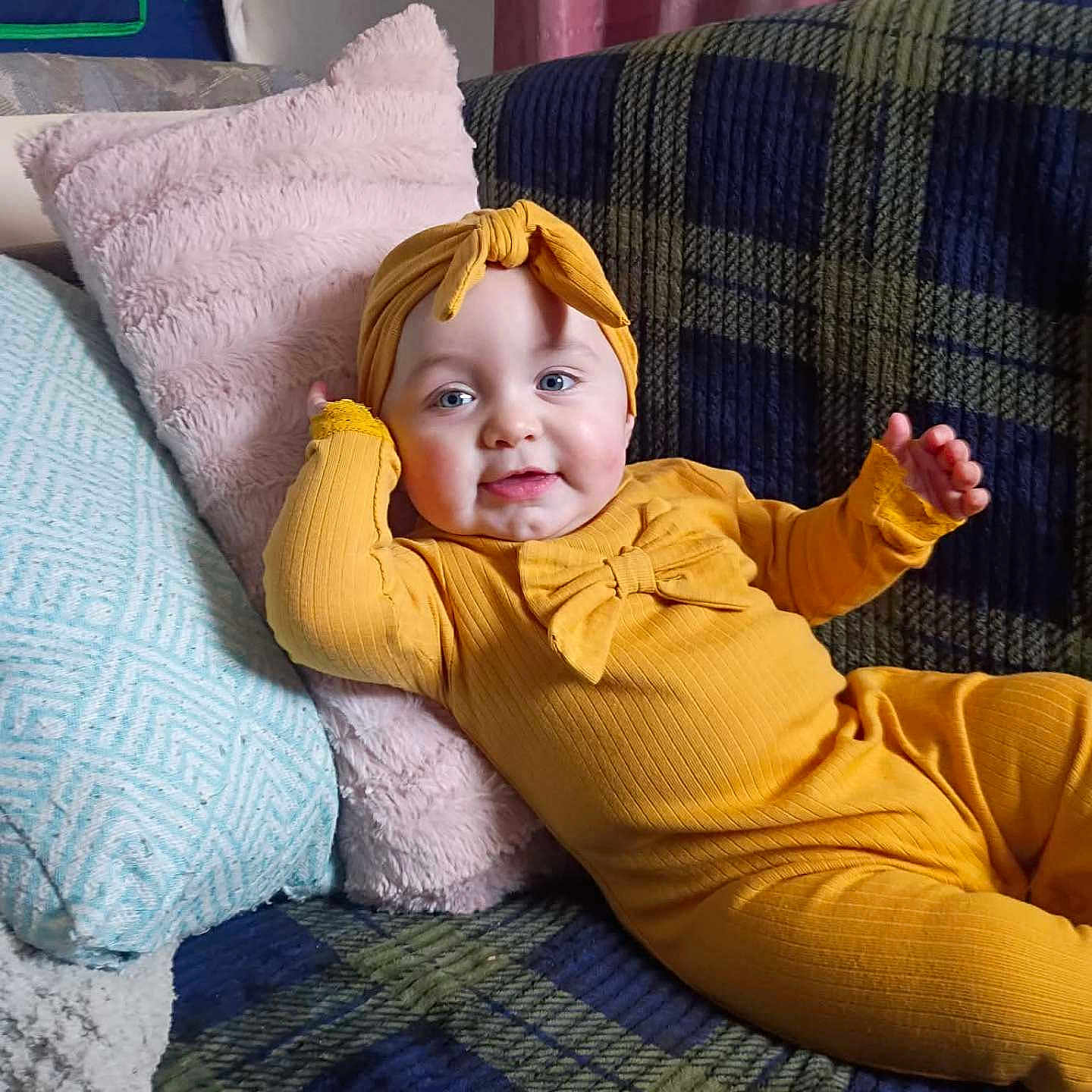 Amelia is registered to the contest to win money with this photo: baby, child, infant, smile, yellow_outfit, headband, couch, pillow, plaid, cozy, indoor, relaxing, cute, portrait, person, furniture, home, soft_texture, happy, comfort