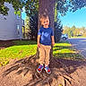 Simon is registered to the contest to win money with this photo: child, boy, smiling, portrait, outdoor, tree, tree_roots, suburban, sunny, daylight, blue_shirt, beige_pants, sneakers, standing, grass, leaves, house, driveway, car, nature