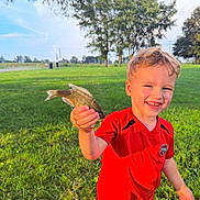 Simon is registered to the contest to win money with this photo: child, boy, fishing, fish, outdoor, park, grass, nature, smiling, happy, portrait, red_shirt, short_hair, summer, sunlight, trees, holding_fish, angler, lake_area, recreation