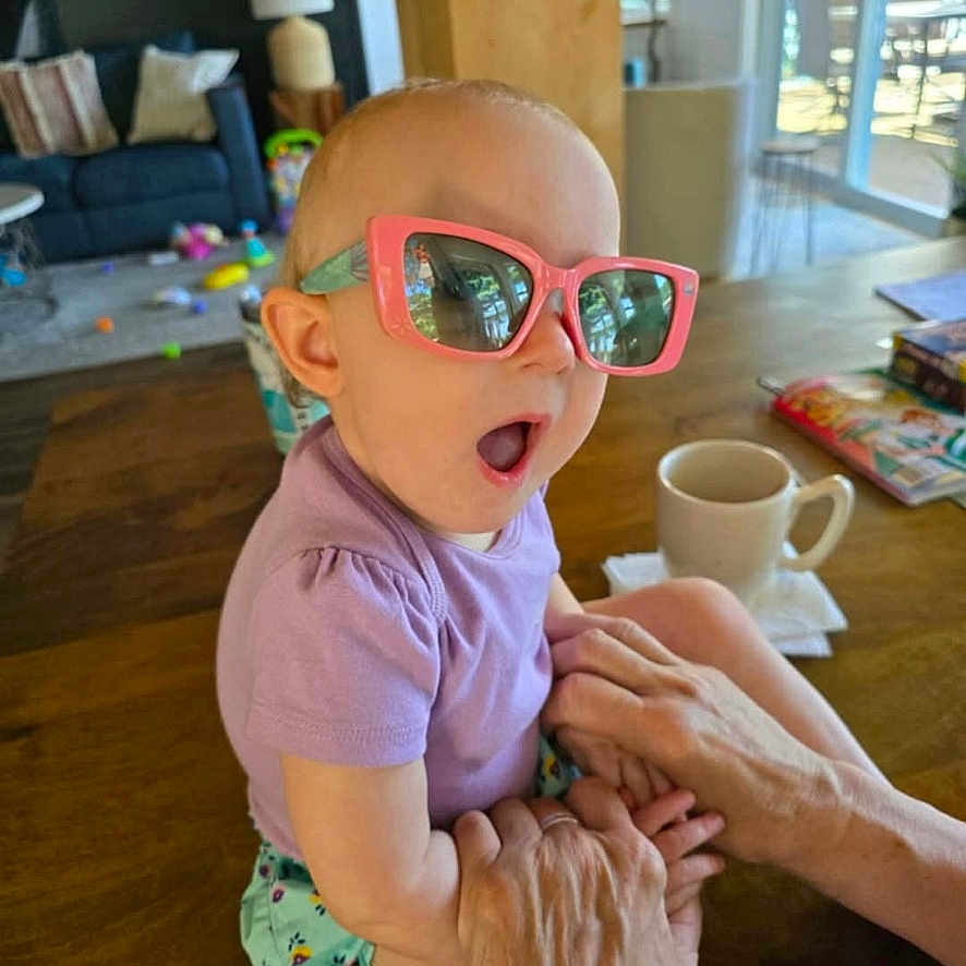 Delilah is registered to the contest to win money with this photo: adult_hand, baby, child, cup, expression, furniture, home, indoor, living_room, person, playful, purple_shirt, reflection, shorts, sunglasses, sunlight, table, toy, window, wood