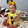 Isabella is registered to the contest to win money with this photo: baby, bed, bedroom, blanket, bow, child, dress, face, family_photo, floral_bedding, happy, headband, indoor, pillow, portrait, remote_control, shoes, sitting, smiling, yellow_outfit