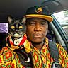 man, cat, car_interior, colorful_clothing, hat, shoulder_pet, black_cat, yellow_eyes, medallion, hoodie, portrait, seatbelt, window, daylight, facial_expression, fashion, pet_accessory, indoor, closeup, person