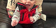 Anthony is registered to the contest to win money with this photo: apparel, baby, black_pants, child, comfort, cozy, cute, fuzzy_slippers, happy, home, indoors, infant, leather_couch, person, plaid_pillow, portrait, red_jacket, sitting, smiling, warm