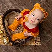Luna is registered to the contest to win money with this photo: baby, child, knitted_clothing, orange, red, bear_ears, heart_shaped_bowl, wooden_floor, honey_toys, blue_eyes, smiling, cute, cozy, portrait, indoors, floor_mat, hand_knit, sitting, infant, looking_up