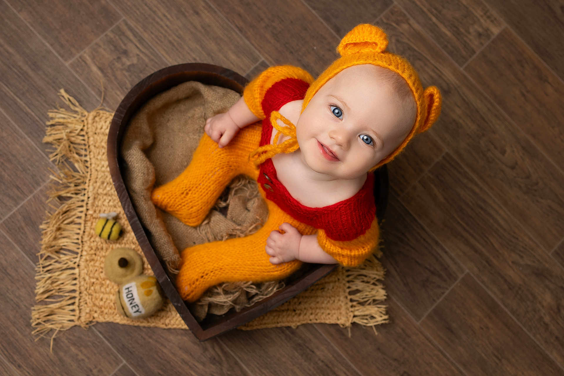 Luna is registered to the contest to win money with this photo: baby, child, knitted_clothing, orange, red, bear_ears, heart_shaped_bowl, wooden_floor, honey_toys, blue_eyes, smiling, cute, cozy, portrait, indoors, floor_mat, hand_knit, sitting, infant, looking_up
