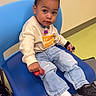 child, toddler, baby, portrait, sitting, chair, blue_chair, waiting_room, indoor, smiling, serious_expression, face, eyes, clothing, sweatshirt, jeans, sneakers, toy, colorful_floor, casual_fashion
