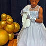 child, girl, dress, tiara, gloves, balloons, gold, white_shawl, wooden_steps, formal_wear, portrait, indoor, event, celebration, fancy, elegant, person, young, fashion, cute