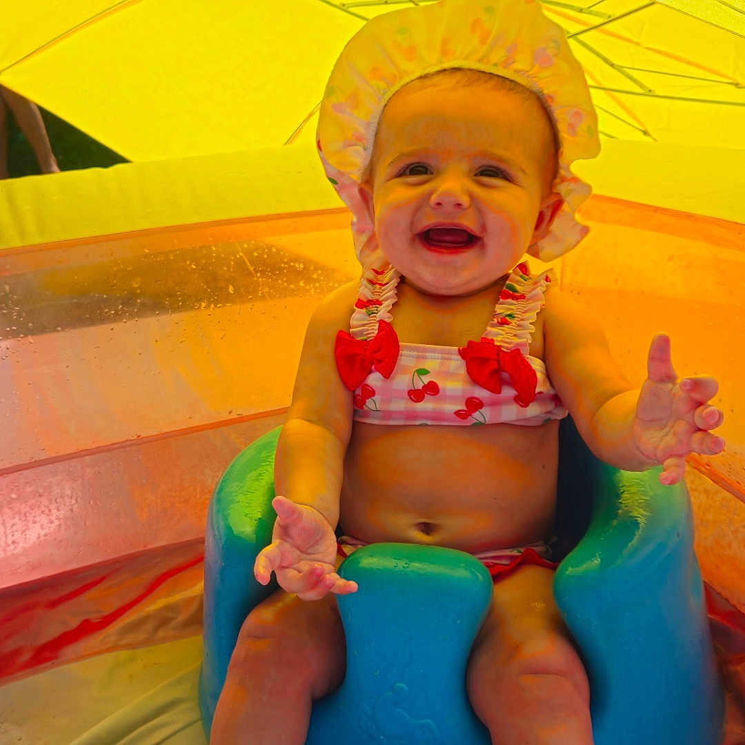 Cassandra Mosier is registered to the contest to win money with this photo: baby, child, smiling, swimsuit, bonnet, blue_seat, pool, water, umbrella, yellow, summer, outdoor, happy, sitting, playful, cute, infant, legs, feet, sunlight