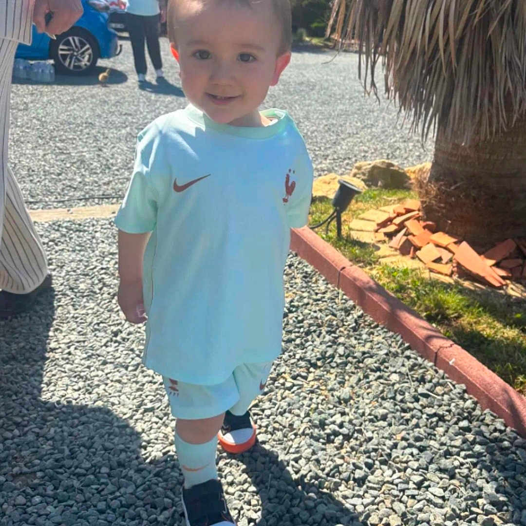 Alone a rejoint le concours — aidez-le/la à gagner de superbes lots ! toddler, child, outdoor, sunlight, gravel, sporty_clothing, sneakers, shadow, palm_tree, car, adult, people, walking, shorts, tshirt, smiling, daytime, nature, sidewalk, casual