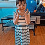 child, girl, striped_dress, barefoot, wooden_floor, living_room, table, television, smiling, happy, indoor, furniture, person, flooring, casual_clothing, home, standing, hands, young, playful