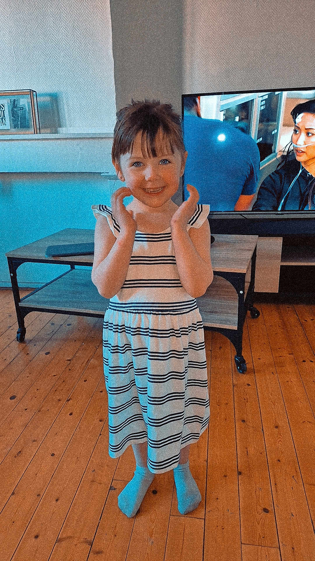 Chelsea participe au concours pour gagner de l'argent avec cette photo : child, girl, striped_dress, barefoot, wooden_floor, living_room, table, television, smiling, happy, indoor, furniture, person, flooring, casual_clothing, home, standing, hands, young, playful
