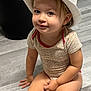 Julia a rejoint le concours — aidez-le/la à gagner de superbes lots ! toddler, child, hat, white_hat, yellow_band, onesie, patterned_clothing, floor, wooden_floor, indoor, smiling, sitting, hands, legs, face, eyes, hair, baby, cute, home