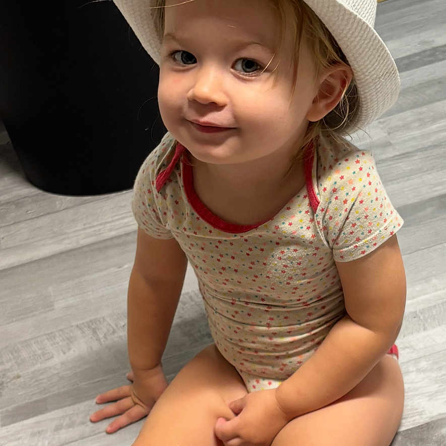 Julia a rejoint le concours — aidez-le/la à gagner de superbes lots ! baby, child, cute, eyes, face, floor, hair, hands, hat, home, indoor, legs, onesie, patterned_clothing, sitting, smiling, toddler, white_hat, wooden_floor, yellow_band