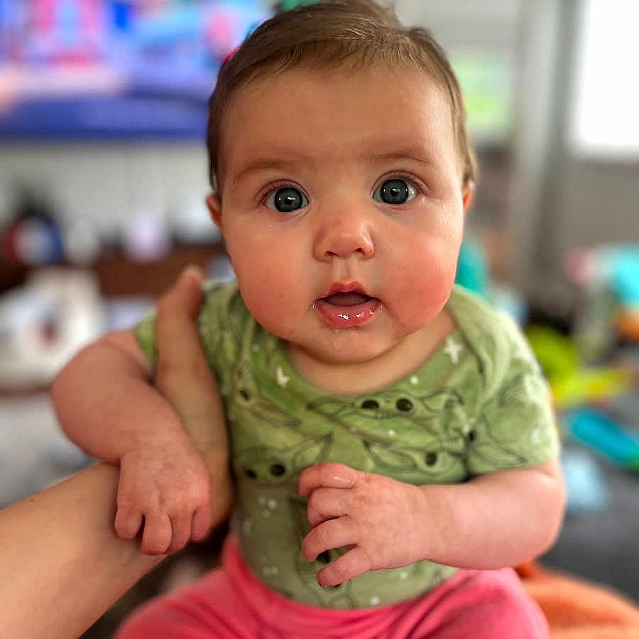 Margie joined the competition — help win amazing prizes! baby, child, clothing, curious, cute, expression, face, green_onesie, hand, head, indoors, infant, living_room, person, pink_pants, portrait, sitting, skin, wide_eyes, young