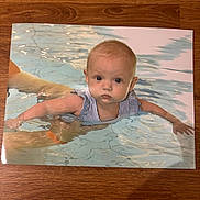 Margot is registered to the contest to win money with this photo: adult_hand, arms, baby, blue, child, expression, face, infant, outdoor, person, pool, reflection, skin, splash, summer, swimming, swimsuit, water, water_surface, young