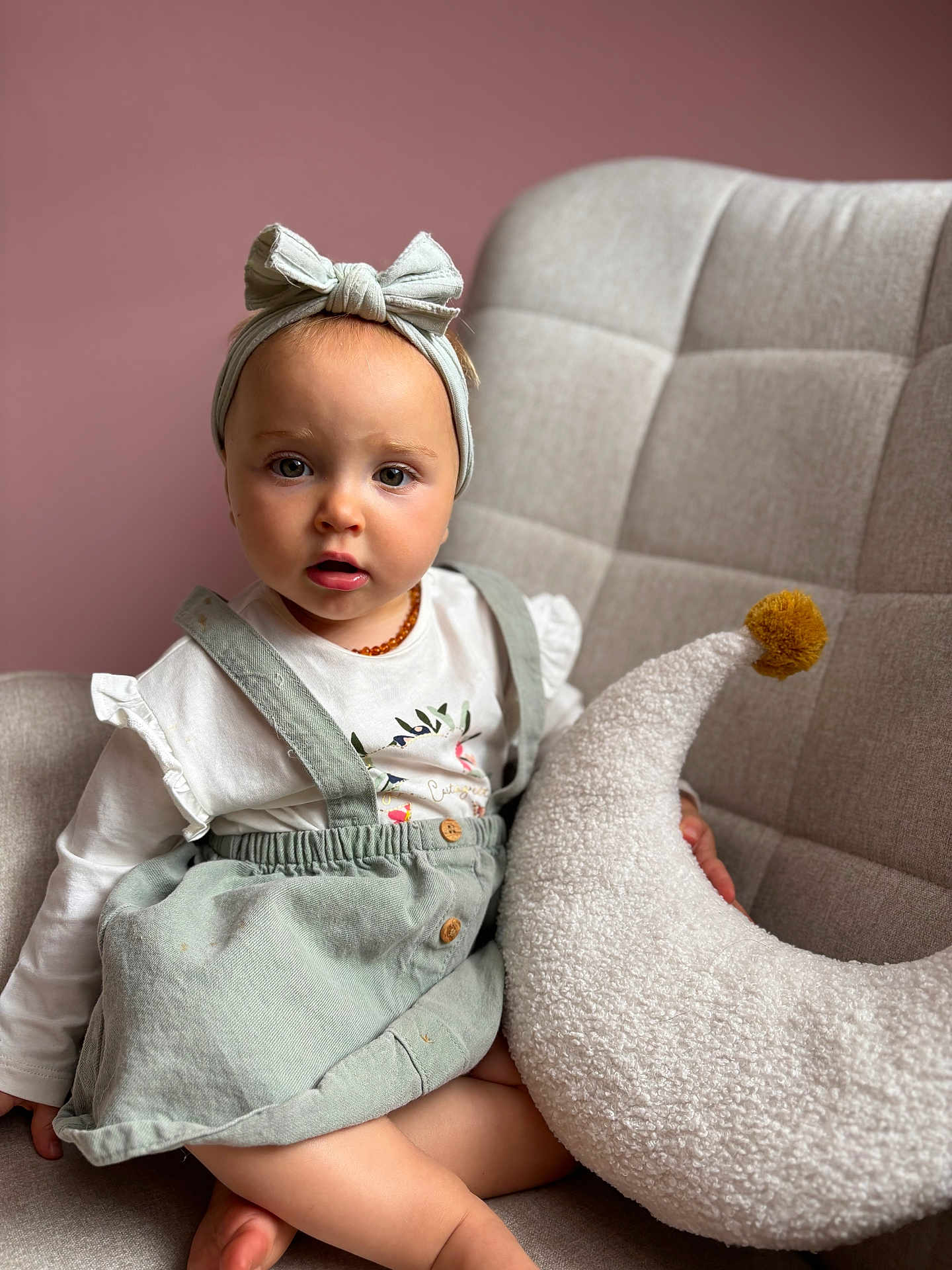 Mència participe au concours pour gagner de l'argent avec cette photo : toddler, child, headband, dress, pillow, crescent_moon, pom_pom, chair, cushion, pink_wall, indoor, sitting, curious, portrait, baby, clothing, soft_texture, person, cute, furniture