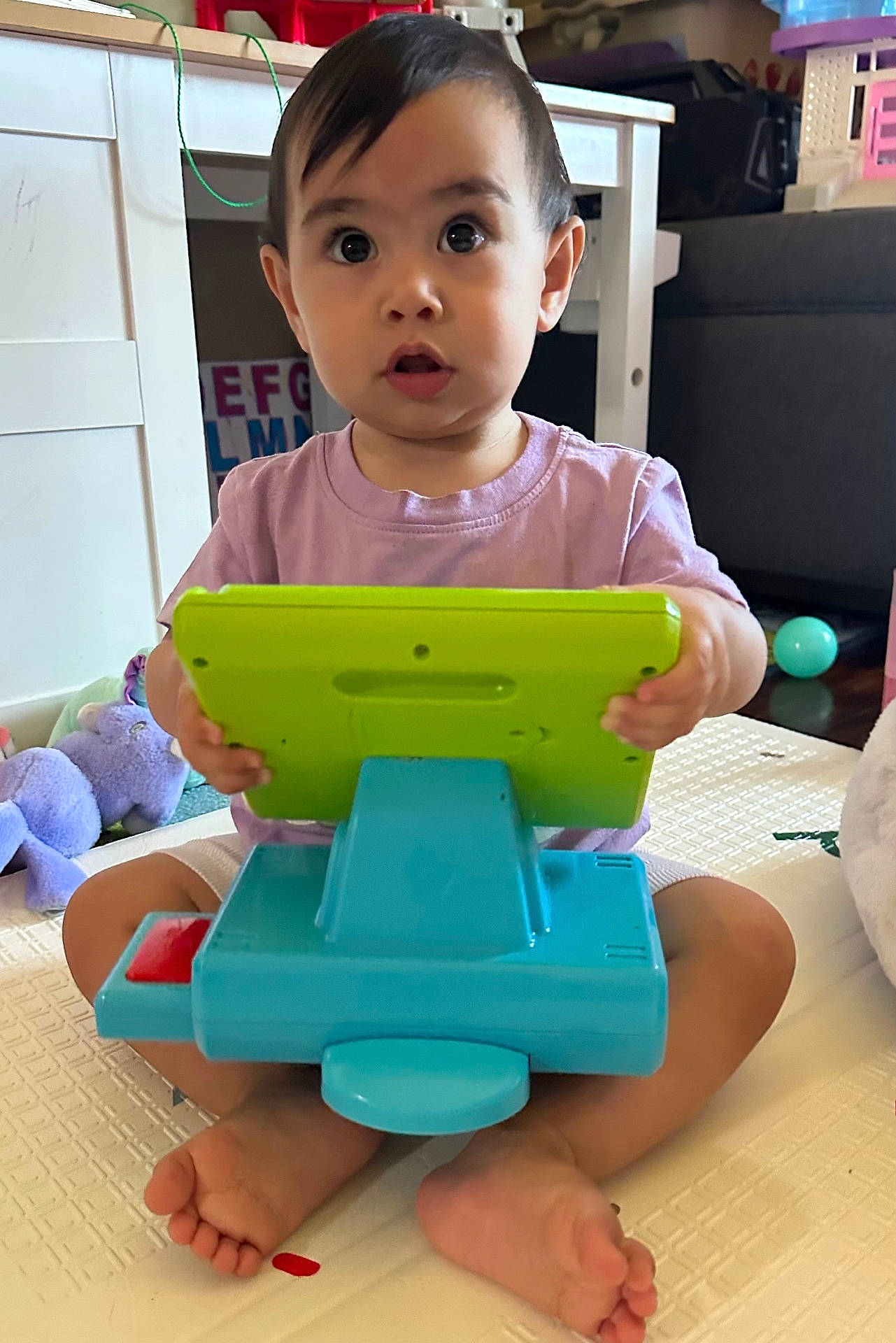 baby, barefoot, cash_register, child, colorful, concentration, expression, face, floor, furniture, home, indoor, person, plastic_toy, play_mat, purple_shirt, sitting, toddler, toy, young_child