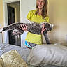 cat, person, smiling, indoors, bedroom, bed, blanket, pillow, door, yellow_shirt, long_hair, portrait, pet, fluffy, gray_fur, holding, standing, home, room, funny