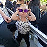 child, toddler, sunglasses, purple_glasses, standing, outdoor, sunlight, crowd, adults, seated_people, portrait, fashion, dress, leopard_print, hair_bow, playful, funny, railings, chairs, park