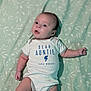 Elijah is registered to the contest to win money with this photo: baby, infant, onesie, text_on_clothing, lying_down, blanket, green, leaf_pattern, cute, expression, small_hands, small_feet, indoor, portrait, child, head, face, eyes, person, soft_lighting
