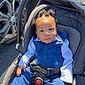 baby, child, stroller, car_wheel, blue_suit, polka_dot_shirt, hair_buns, seatbelt, outdoor, sunlight, curious_expression, infant, car_seat, toddler, parking_lot, person, casual, cute, portrait, young