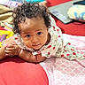 baby, curly_hair, red_surface, dress, strawberry_pattern, blanket, laptop, indoor, child, cute, gaze, expression, infant, lying_down, homely, soft_lighting, portrait, closeup, comfortable, cozy