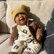 Jett joined the competition — help win amazing prizes! baby, child, smiling, pajamas, hat, pom_poms, couch, beige, brown, cream, bear_pattern, clothing, indoor, cozy, happy, person, sitting, cute, infant, portrait